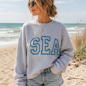 May include: A light gray sweatshirt with the words "Vitamin SEA" embroidered in blue. The sweatshirt is worn with denim shorts on a sunny day at the beach. The person is wearing sunglasses and a necklace.