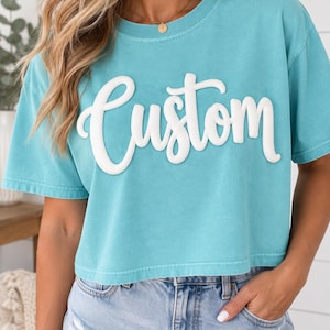 May include: A turquoise short-sleeved crop top featuring the word Custom in large, raised, white, three-dimensional script lettering across the chest. The garment has a crew neckline and a relaxed fit with a straight hemline at the waist.