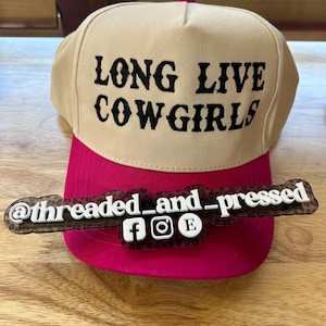 Op de afbeelding: Een beige en fuchsia baseballpet met de tekst "LONG LIVE COWGIRLS" in zwart. De pet heeft een fuchsia klep en een decoratief label met de tekst "@threaded_and_pressed" en social media-iconen.