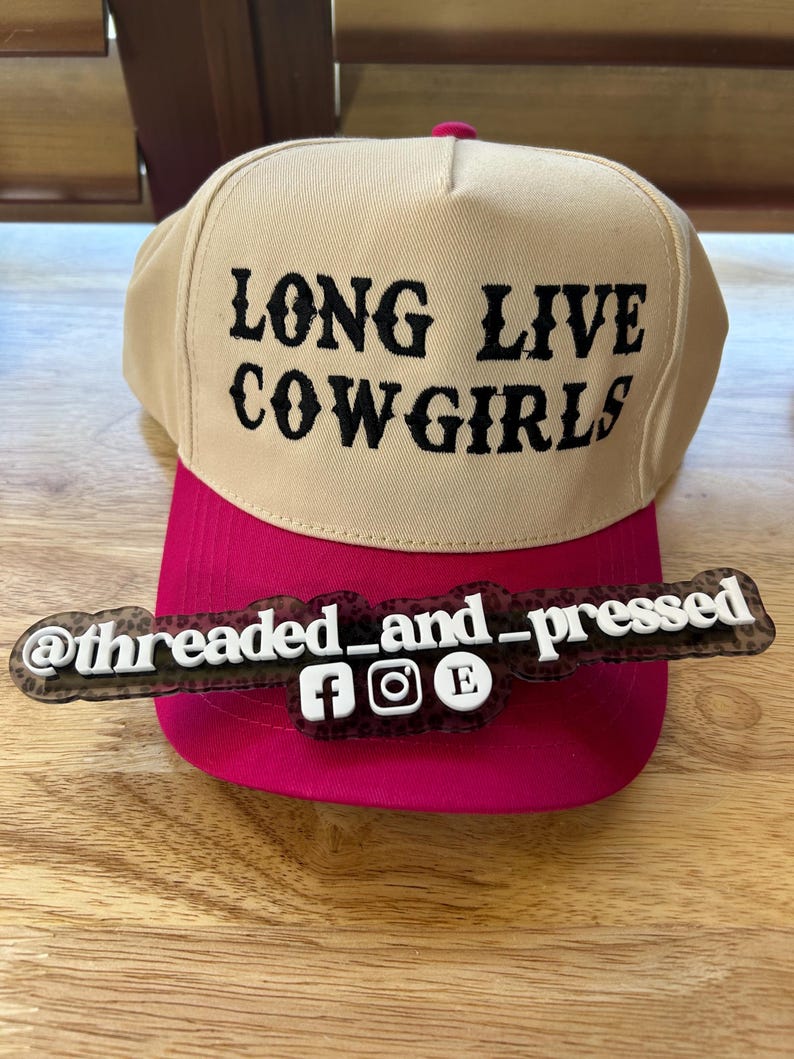 Op de afbeelding: Een beige en fuchsia baseballpet met de tekst "LONG LIVE COWGIRLS" in zwart. De pet heeft een fuchsia klep en een decoratief label met de tekst "@threaded_and_pressed" en social media-iconen.