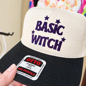 May include: A two-tone baseball cap with a cream-colored front panel and a black brim. The front features the purple text "BASIC WITCH" with four small purple stars. A sticker on the brim reads "OTTO PREMIUM FABRIC CLASSIC FIT OSFM MID PROFILE".
