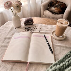 Puede incluir: Un diario abierto con el texto "Dear Little One" está sobre una mesa, junto a un bolígrafo, un jarrón con flores y un vaso de café helado. Un gato está acurrucado en una silla en el fondo. Una escena acogedora y relajante.