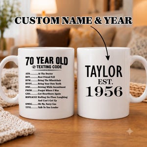 May include: Two white ceramic mugs. One mug reads "70 YEAR OLD TEXTING CODE" with a list of abbreviations and their meanings. The other mug says "TAYLOR EST. 1956". The mugs are on a table.