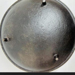 Pode incluir: Uma panela redonda de ferro fundido preto com três pernas curtas e resistentes na parte inferior. A superfície tem um acabamento fosco e texturizado. O texto em relevo na base diz "GRISWOLD" em um arco acima de "ERIE" e "PA" com o número "8".
