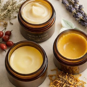 May include: Three open amber glass jars filled with creamy, light-colored balm. The jars are labeled "Sacred Grove" and "Acne Calm Balm." Dried herbs, rose hips, and lavender sprigs surround the jars, suggesting natural ingredients.