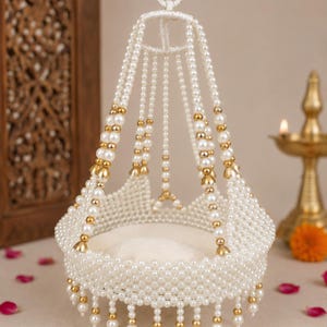 May include: A decorative hanging cradle made of white faux pearls and gold-toned metal beads. The structure features a circular base with a beaded fringe and multiple beaded strands extending upward to a central ring for suspension.