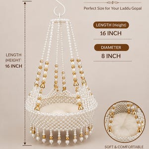 May include: A hanging swing for a Laddu Gopal idol, crafted from white faux pearls and gold-toned beads. It features a circular base with a soft white cushion, measuring 16 inches in height and 8 inches in diameter, suspended by a white metal S-hook.