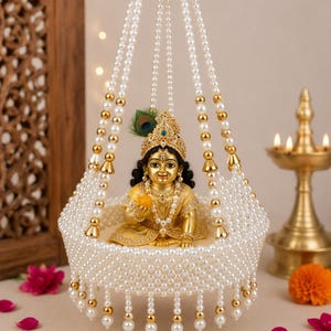 May include: A golden figurine of a child deity seated inside a hanging cradle constructed from white faux pearls and gold-toned beads. The figure wears an ornate crown with a peacock feather and holds a small yellow object, adorned with jewelry.