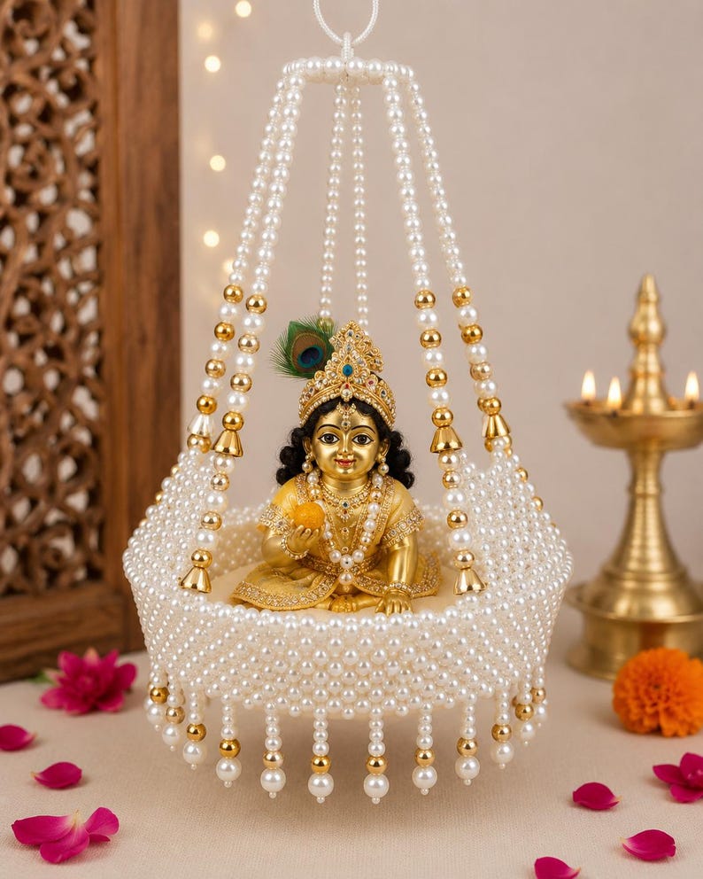 May include: A golden figurine of a child deity seated inside a hanging cradle constructed from white faux pearls and gold-toned beads. The figure wears an ornate crown with a peacock feather and holds a small yellow object, adorned with jewelry.
