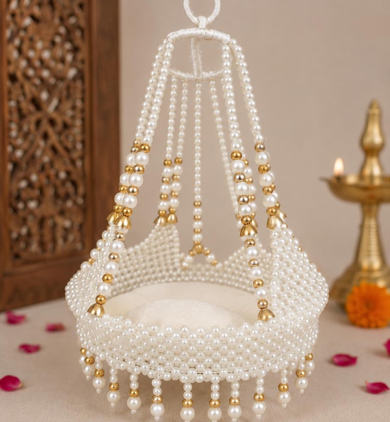 May include: A decorative hanging cradle made of white faux pearls and gold-toned metal beads. The structure features a circular base with a beaded fringe and multiple beaded strands extending upward to a central ring for suspension.