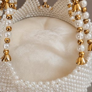 May include: A decorative circular object featuring a central off-white fabric cushion surrounded by a woven structure of white faux pearls and metallic gold-toned beads. The design includes hanging beaded strands with gold-toned bell-shaped accents.