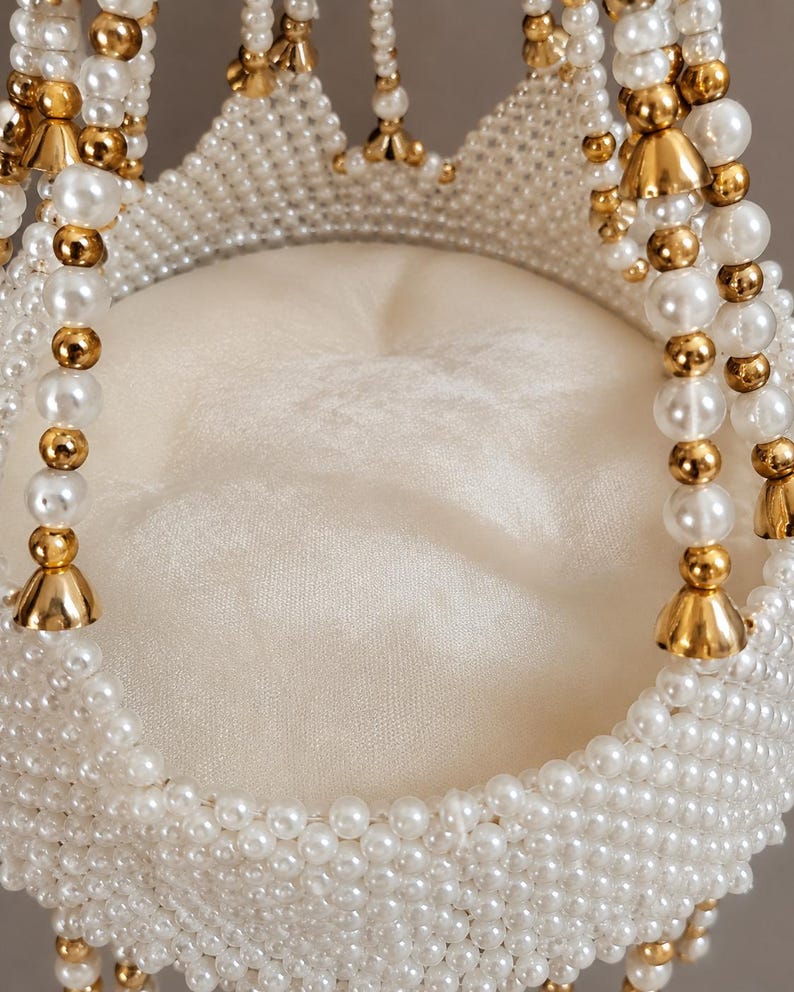 May include: A decorative circular object featuring a central off-white fabric cushion surrounded by a woven structure of white faux pearls and metallic gold-toned beads. The design includes hanging beaded strands with gold-toned bell-shaped accents.