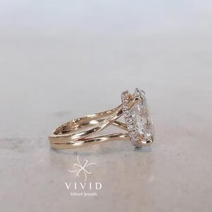 Puede incluir: Un anillo de oro rosa con una gran piedra central de corte ovalado rodeada por un halo de pequeños diamantes redondos. La banda tiene un diseño de vástago dividido que se estrecha hacia el engaste. El texto VIVID Velvet Jewels aparece debajo del anillo.