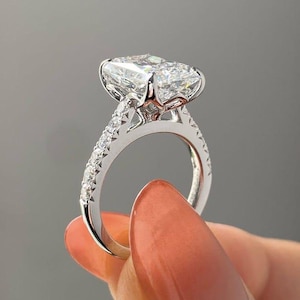 May include: A platinum engagement ring with a large, rectangular-cut diamond. The central stone is held by prongs, and the band is adorned with smaller diamonds. The ring is displayed against a grey backdrop, showcasing its sparkle and design.