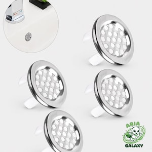 May include: Four stainless steel sink strainers with white plastic tabs. Each strainer has a perforated centre and a shiny, metallic finish. The image also shows a sink and tap in the background.