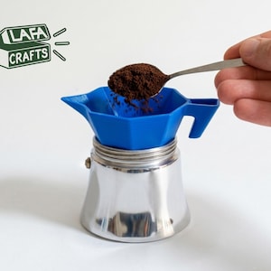 Può includere: Una base per caffettiera moka in metallo sormontata da un imbuto in plastica blu brillante. Un cucchiaio d'argento versa caffè macinato marrone scuro nell'imbuto. Il logo LAFA CRAFTS è visibile in alto a sinistra in verde scuro.