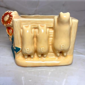 May include: A cream-colored ceramic planter shaped like a wooden fence with three small pigs leaning over the top. The side of the planter features a painted sunflower with orange petals and green leaves. The planter has a hollow center for holding items.