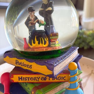 May include: A snow globe featuring two figures standing by a cauldron, resting on a stack of books labeled Potions and History of Magic. The base includes a blue potion bottle with a yellow spiral and a red flask. The globe contains liquid.