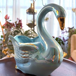 Peut inclure: Un cache-pot en céramique en forme de cygne bleu clair avec une finition irisée et des accents peints en or sur le bec et les plumes des ailes. Le cygne a un long cou courbé et un corps creux conçu pour contenir des objets ou des plantes.