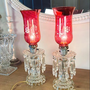 May include: A pair of electric lamps featuring red glass hurricane shades with etched floral patterns. The clear glass bases are adorned with hanging crystal prisms and a scalloped design. Each lamp includes a light bulb and a power cord.