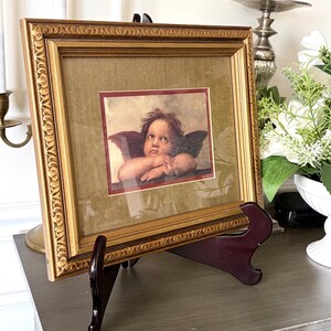 May include: A rectangular gold-toned picture frame with an ornate, textured border containing a print of a cherub with wings, resting its chin on its hands. The print is set against a dark olive green mat with a thin red inner border.