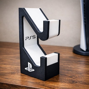 May include: A black and white PS5 console stand. The stand has a textured black frame with white interior panels. The PS5 logo is visible on the front. The stand is designed to hold a PS5 console.
