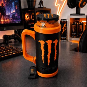 Könnte beinhalten: Ein schwarzer und orangefarbener Getränkedosenhalter mit Griff, der das Monster Energy-Logo in Orange auf der Vorderseite zeigt. Der Halter enthält ein kleines schwarzes Etikett mit demselben Logo an einer Kette und eine Monster Energy Taurine Voodoo Dose.