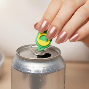 Puede incluir: Primer plano de una lata de aluminio plateada con condensación. Se utiliza un abrelatas verde y amarillo para levantar la pestaña. El abrelatas está diseñado para facilitar la apertura de latas.