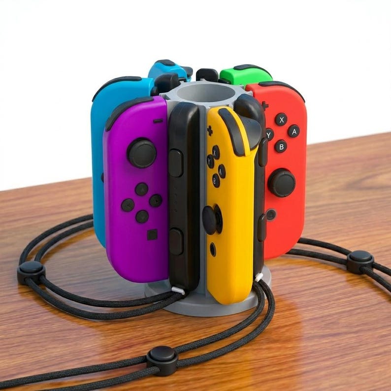 May include: A multi-colored game controller holder with controllers in blue, purple, black, yellow, red, and green. The holder is gray and circular, with a black cord around the base. The controllers are arranged around the holder.