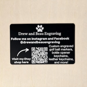 May include: A rectangular black business card featuring a white paw print icon and the text Drew and Beau Engraving. It includes social media handles, a QR code with an arrow pointing to it, and text advertising custom engraved golf ball markers.