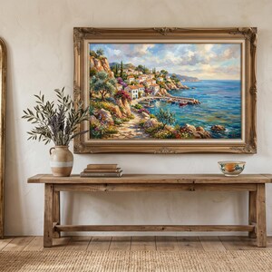 Puede incluir: Una pintura enmarcada de un paisaje de un pueblo costero cuelga sobre una consola rústica de madera. La obra de arte muestra un camino, edificios y un muelle. Un gran espejo con marco dorado y un sillón beige también están en la habitación.