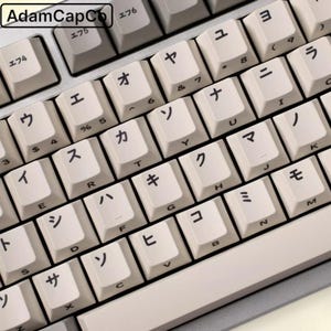 Op de afbeelding: Een mechanisch toetsenbord met gebroken witte toetsen voorzien van zwarte Japanse katakana-tekens en standaard Engelse letters. In de linkerbovenhoek staat de tekst AdamCapCo. Het frame van het toetsenbord heeft een neutrale grijze kleur.