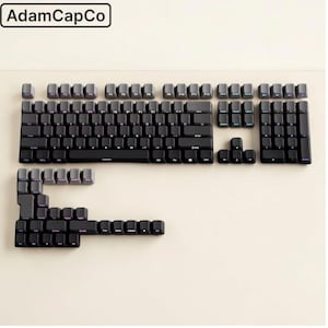 May include: A set of black and dark grey mechanical keyboard keycaps arranged in a layout. The set includes a main alphanumeric block, a number pad, function keys, and navigation keys. The text AdamCapCo is visible in the top left corner.