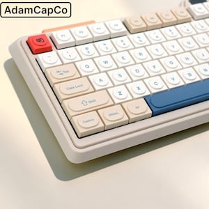 May include: A mechanical keyboard with a beige frame and round white keycaps featuring blue lettering. The layout includes a red escape key, beige modifier keys, and a blue spacebar. One key is labelled 250mL. The text AdamCapCo is visible.