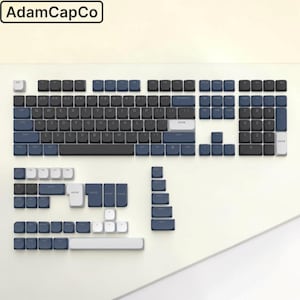 Op de afbeelding: Een mechanische toetsenbord-keycapset met een kleurenschema van donkerblauwe, zwarte en witte toetsen. De lay-out bevat een volledige QWERTY-indeling met numeriek toetsenbord, plus extra losse toetsen in verschillende maten. De tekst AdamCapCo is zichtbaar.