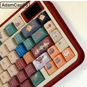 Può includere: Una tastiera meccanica con tasti personalizzati raffiguranti illustrazioni di personaggi, gufi, lettere e simboli magici. La tastiera ha una cornice rosso scuro e una base color crema. Il testo AdamCapCo è visibile nell'angolo in alto a sinistra.