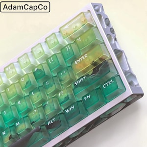 May include: A mechanical keyboard with translucent green and yellow gradient keycaps featuring white lettering. The keyboard has a silver metallic frame with a geometric cutout pattern along the side. The text AdamCapCo is visible in the corner.
