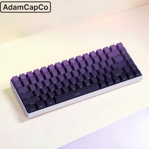 May include: A mechanical keyboard with a white frame and a gradient keycap set that transitions from light purple at the top to dark purple at the bottom. The keys feature white lettering. The text AdamCapCo is visible in the top left corner.