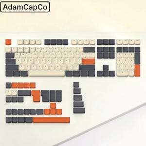 May include: A mechanical keyboard keycap set featuring a color scheme of cream, dark gray, and orange. The set includes a full QWERTY layout with a number pad, plus additional loose keycaps in various sizes and colors. The text AdamCapCo is visible.