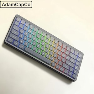 May include: A black mechanical keyboard with rounded, translucent keycaps featuring white lettering. The keyboard displays a vibrant rainbow-coloured LED backlight glowing from beneath the keys. The text AdamCapCo is visible in the top left corner.