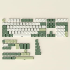 Könnte beinhalten: Ein mechanisches Tastatur-Tastenkappen-Set in einer Farbpalette aus Weiß, Salbeigrün und Olivgrün. Das Set enthält ein vollständiges QWERTY-Layout, einen Ziffernblock und verschiedene alternative Tasten, die flach angeordnet sind.