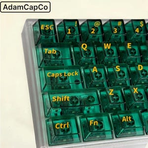 May include: A mechanical keyboard featuring translucent dark green keycaps with bright yellow lettering. The keys display standard QWERTY layout characters including ESC, Tab, Caps Lock, Shift, Ctrl, Fn, and Alt. The brand name AdamCapCo is visible.