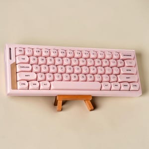 Peut inclure: Un clavier mécanique rose clair doté de touches rondes de style machine à écrire avec des lettres noires. Le clavier présente un cadre rectangulaire avec un panneau d'accentuation en or métallique sur le côté gauche et une longue barre d'espace.