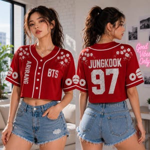 May include: A red cropped baseball jersey featuring white trim, white floral embroidery on the shoulders, and white text reading ARIRANG and BTS on the front. The back displays the name JUNGKOOK and the number 97 in large white glittery lettering.