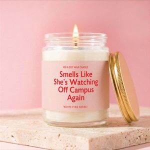 May include: A clear glass jar candle with a lit wick and a gold metal lid. The label is light pink with red text that reads: 100% SOY WAX CANDLE, Smells Like She's Watching Off Campus Again, WHITE PINE FOREST. The candle contains white wax.