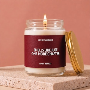 May include: A lit soy wax candle in a clear glass jar with a gold-colored metal lid. The label is dark red with white text that reads 100% SOY WAX CANDLE, SMELLS LIKE JUST ONE MORE CHAPTER, and BEACH RETREAT. The candle has a single burning wick.