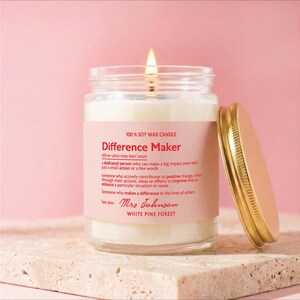 May include: A lit soy wax candle in a clear glass jar with a gold metal lid. The label features the text Difference Maker, a dictionary-style definition, the name Mrs Johnson, and the scent White Pine Forest printed in red on a pink background.
