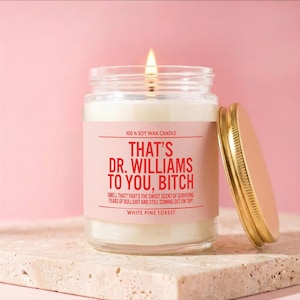 May include: A lit soy wax candle in a glass jar with a gold lid. The label reads "THAT'S DR. WILLIAMS TO YOU, BITCH" in red text. The candle is on a stone surface against a pink background. The text also says "White Pine Forest".
