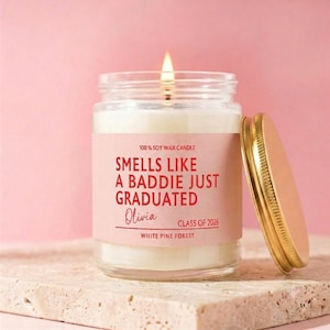 May include: A clear glass jar candle with a lit wick and a gold metal lid. The label is light pink with red text that reads: 100% SOY WAX CANDLE, SMELLS LIKE A BADDIE JUST GRADUATED, Olivia, CLASS OF 2026, WHITE PINE FOREST.