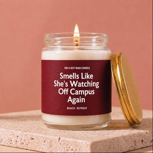 May include: A lit soy wax candle in a clear glass jar with a gold-colored metal lid. The label is deep red with white text that reads 100% SOY WAX CANDLE, Smells Like She’s Watching Off Campus Again, and BEACH RETREAT in a sans-serif font.